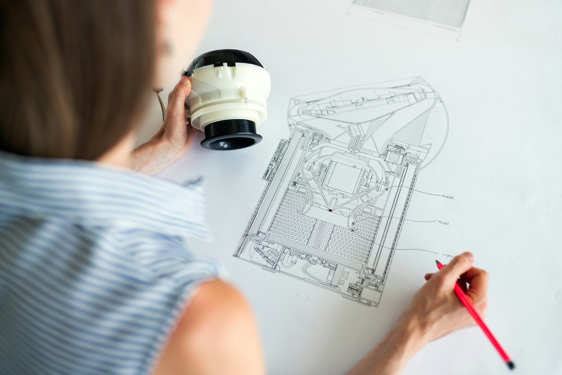 A hand sketching over a technical drawing with a printed part in the other hand